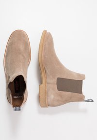 Beige mokkasiner med elastiske sidepaneler, gummisåler og broderidetaljer. Inkluderer en pull-tab for enkel påkledning.
