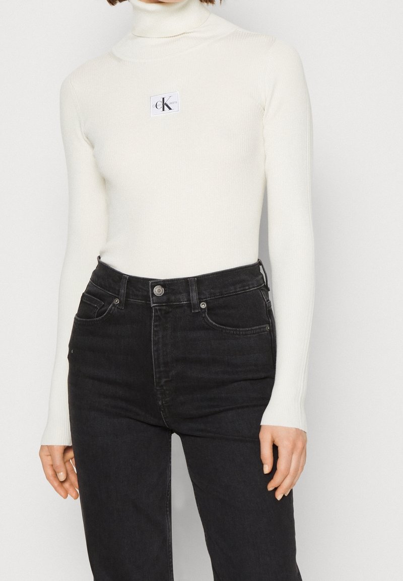 Femme portant un pull col roulé blanc côtelé avec un petit écusson logo Calvin Klein sur la poitrine et un jean noir taille haute.