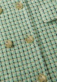 Gros plan sur un tissu en tweed vert et crème avec un tissage texturé et des boutons ronds dorés brillants disposés en rangées.