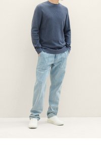 Pull tricoté bleu à col rond, associé à un jean denim ample bleu clair et des baskets blanches, sur un fond neutre.