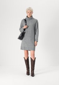 Robe en tricot grise avec col montant côtelé, arborant un motif en losange. Assortie à des bottes de cow-boy en cuir marron et un sac à main noir.