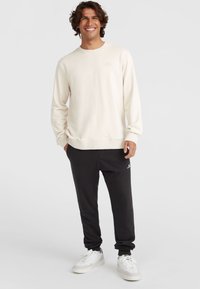 Lichtgekleurde cremewitte sweater met ronde hals en ribgebreide boorden, gecombineerd met zwarte tapered joggers en witte sportieve schoenen, met een logo-accent.
