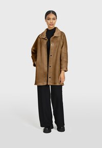 Veste en fausse fourrure marron avec un col, des boutons noirs et une coupe décontractée. Associée à un pantalon large noir et des chaussures à plateforme.