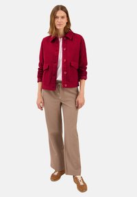 Femme se tenant debout, portant une veste texturée rouge à boutons, un haut blanc, un pantalon beige à cordon de serrage et des baskets marron sur un fond uni.