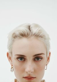 Persona con cabello corto platino, piel clara y ojos azules, vistiendo aretes geométricos de aro en plata y oro que no coinciden, sobre un fondo blanco liso.