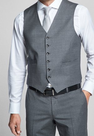 Homme portant une chemise blanche, un gilet gris boutonné, un pantalon gris assorti, une ceinture noire et une cravate claire à motifs, debout avec une main dans la poche.