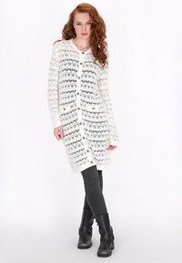 Bílý pletený kardigan s krajkovým vzorem, předními knoflíky, dlouhými rukávy a rovný fit, kombinovaný s tmavě šedými skinny džínami a černými kotníkovými botami.