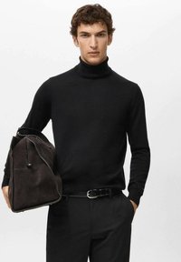 Zwarte coltrui van fijngebreid materiaal, gecombineerd met donkere broek. De persoon houdt een bruine suède tas met rits vast.