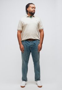 Άνδρας με πλεγμένα μαλλιά, ντυμένος με μπεζ πόλο πουκάμισο, μπλε τζιν, λευκά αθλητικά παπούτσια, στέκεται μπροστά σε απλό ανοιχτό φόντο, κοιτάζοντας προς τα πλάγια.