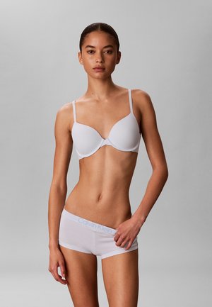 Kvinde med samlet hår, iført hvid bh og matchende Calvin Klein boyshort undertøj, står mod en ensfarvet grå baggrund.