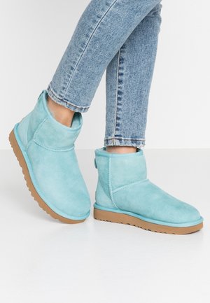 Bottines en daim bleu clair avec semelles en caoutchouc marron portées avec un jean en denim bleu clair sur un fond blanc.