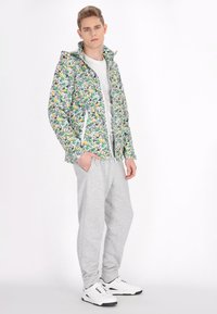 Giacca con zip a motivo floreale in bianco, verde e giallo. Tessuto morbido, stile aderente, abbinata a pantaloni della tuta grigi e sneakers bianche.