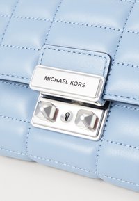 Geantă din piele bleu deschis cu un design matlasat, accesorii din metal argintiu și închidere cu lacăt. Prezintă un plăcuță de marcă metalică și bolțuri proeminente.