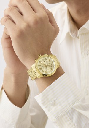 Nærbilde av en person som har på seg et gullfarget metallarmbånd med kronografklokke på venstre håndledd, med hendene samlet nær ansiktet.