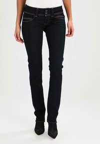 Slim-fit mörkblå jeans med dubbelknäppning, bärs med svarta spetsiga stövlar och en vit topp, visas från midjan till fötterna.