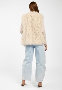 Fake fur mouwloos vest in crème kleur met een harige textuur, gecombineerd met lichtblauwe wijde jeans en neutrale sandalen.