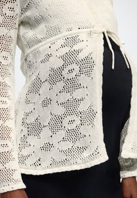 Κοντινό πλάνο ριγωτού πλεκτού πουλόβερ με κεντήματα λουλουδιών πάνω από μαύρο φόρεμα εγκυμοσύνης, παρουσιάζοντας λεπτομέρεια δεμένης μέσης και υφή στο μανίκι.