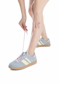 Zapatilla de ante azul claro con acentos rosas, franjas crema y suela de goma. Los cordones están siendo atados, mostrando el diseño del zapato.