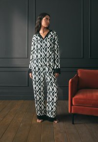 Set de pijama cu model alb-negru, prezentând motive cu mâini, mâneci lungi și manșete contrastante negre, asociat cu un scaun tapițat roșu.