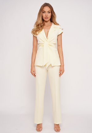 Kobieta o długich, falowanych, blond włosach, ubrana w kremową, krótką bluzkę zapinaną na kopertę i pasujące proste spodnie, stojąca na tle jednolitego białego tła.