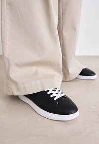 Black leather sneakers with white laces and a white rubber sole, paired with loose beige trousers, on a light textured surface.