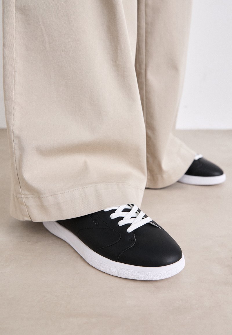 Černé kožené tenisky s bílými tkaničkami a bílou gumovou podrážkou, kombinované s volnými béžovými kalhotami, na světle texturovaném povrchu.