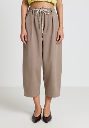 Femme portant un pantalon beige large et court avec un cordon blanc et des talons noirs pointus, vue de la taille aux pieds.