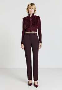 Bordeauxfarbene kurze Jacke mit Kunstfelloberteil und gerippten Ärmeln, kombiniert mit maßgeschneiderten, dunkelroten Hosen und passenden spitzen High Heels.