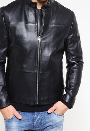 Hombre con una chaqueta de cuero negra con cremallera sobre una camiseta negra, con las manos en los bolsillos de unos vaqueros azul claro.