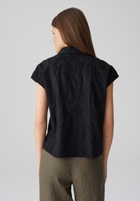 Blusa nera a maniche corte con motivo a foglia testurizzato, dotata di colletto e risvolti, abbinata a pantaloni di lino color khaki.