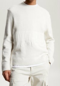 Vit pullovertröja gjord av mjukt, fluffigt material med ribbade ärmslut och nederkant. Subtil strukturerad mönster med en delvis synlig logotyp.