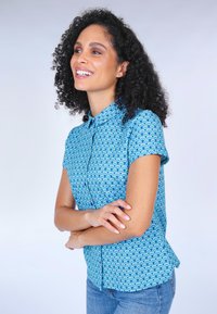 Short-sleeve, collared shirt in blue fabric featuring a wave pattern in lighter blue. The shirt has a relaxed fit and curved hem.