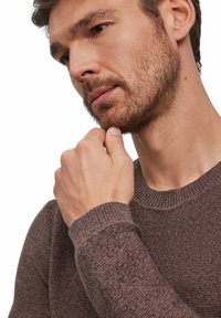 Brauner, strukturierter Pullover mit geripptem Kragen, getragen von einer Person, die ihr Kinn berührt. Die Aufnahme zeigt eine Nahaufnahme der Stoff- und Passformdetails.