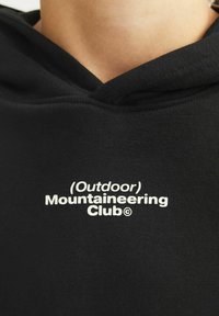 Melnas sporta izdilis ar kapuci, ar baltu drukātu logotipu, kas priekšā lasa "(Āra) Kalnu iepazīšanas klubs". Mīksta auduma tekstūra.