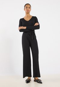 Black jumpsuit with long sleeves, V-neckline, and a tied waistband. Features wide legs and smooth fabric, paired with black pointed flats.