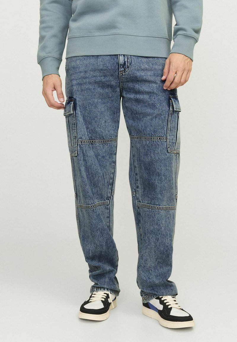 Jeans cargo blu chiaro con molte tasche, vestibilità leggermente ampia e dettagli di cucitura visibili, abbinati a sneakers nere e bianche.