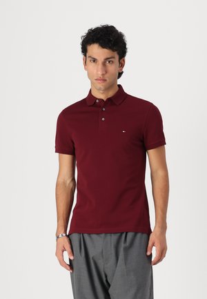 Burgunderfarbenes Poloshirt mit kurzen Ärmeln, einer Drei-Knopf-Leiste und dezentem Logo auf der Brust; kombiniert mit grauen Hosen.