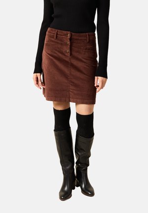 Vrouw met een bruine ribfluwelen rok met knopen aan de voorkant, een zwart shirt met lange mouwen, zwarte kniekousen en zwarte leren laarzen.