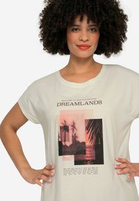Camiseta de manga corta en color beige claro con un estampado gráfico de un atardecer y palmeras, acompañado de texto en marrón oscuro. Textura suave.