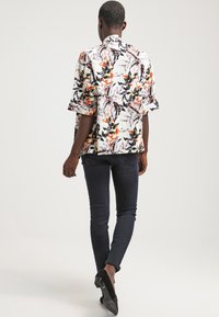 Košile s krátkým rukávem s živým květinovým vzorem v červené, oranžové a černé na světlém pozadí; doplněná tmavými přiléhavými džíny.
