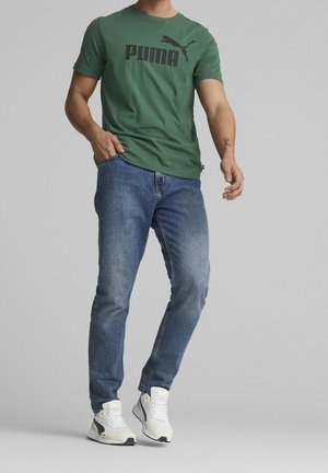 Homme portant un t-shirt vert Puma, un jean bleu et des baskets blanches, debout devant un fond gris uni.
