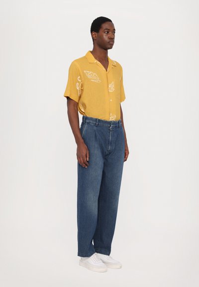 Kortermet gul skjorte med hvit trykk, paret med løse blå denimbukser og hvite sneakers. Enkelt design, avslappet passform, ingen synlige detaljer.