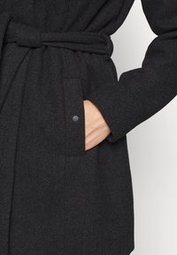 Manteau en laine gris foncé avec ceinture, poches latérales et manches longues, main partiellement dans la poche sur un fond clair uni.