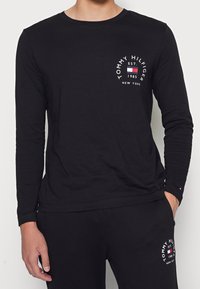 Mustat pitkät hihaiset T-paidat, joissa on puuvillakangas, sisältävät ympyrälokin, jossa on "TOMMY HILFIGER" -teksti, perustamispäivämäärä ja korostusvärit rinnassa.