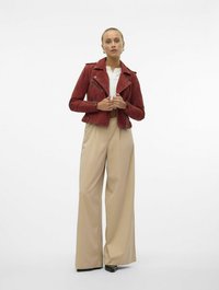 Rode suède motorjack met ritszakken en een cropped pasvorm, gecombineerd met beige wijde broek en zwarte schoenen. Witte blouse met kraag eronder.