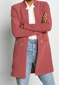 Světle růžový blazer s žebrovanou texturou, dvěma předními kapsami se zipem a volným střihem. Nositý otevřený přes bílou košili a džíny.