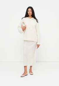 Kremfarget, teksturert oversized hettegenser med snorer, sammen med et hvitt, langt skjørt og myke rosa leppestifter, holder en lysbeige veske.