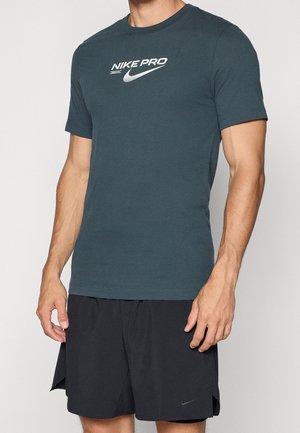 Mann trägt ein dunkelgrünes Nike Pro Kurzarmshirt und schwarze Nike Sportshorts, steht mit entspannt an den Seiten hängenden Armen.