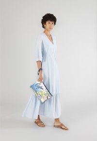 Ljusblå, långärmad klänning med en rynkad kjol, bärs med bruna sandaler. Håller en färgglad handväska med blommönster.