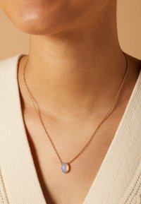 Gouden ketting met een ovale hanger waarin een gladde, iriserende blauwe steen is gezet in een gestructureerd, rond montuur.